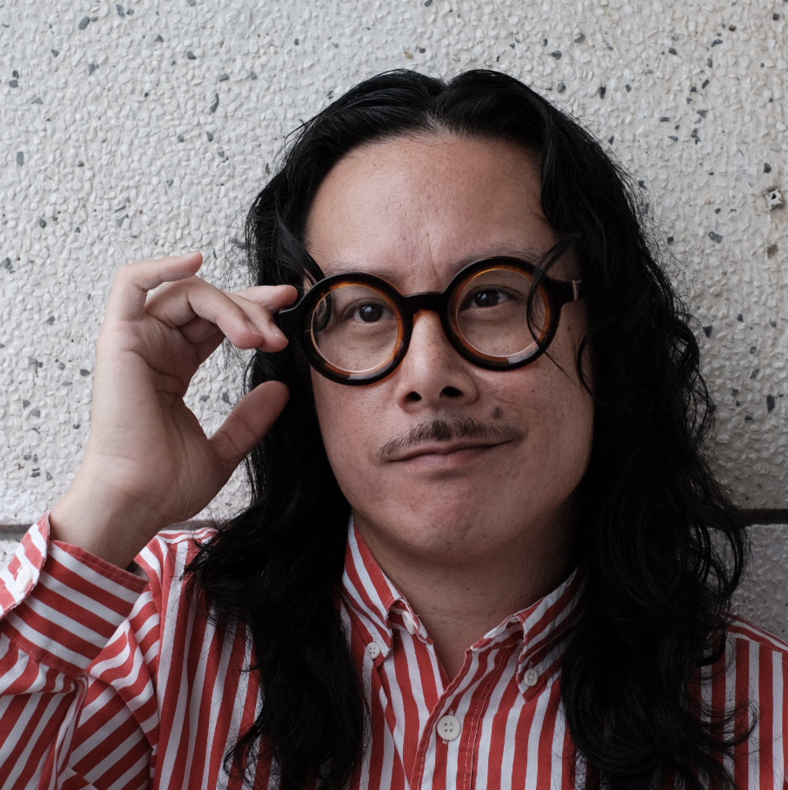 Jimmy Luu, lifting a pair of round tortoiseshell glasses, against a pebbled concrete wall.