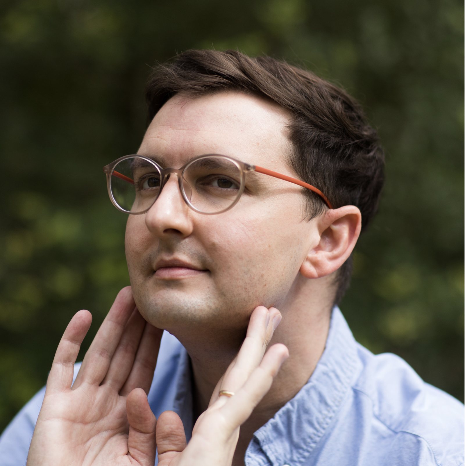 Ryan Menefee, hands framing his jaw, wearing pale round glasses against green foliage.